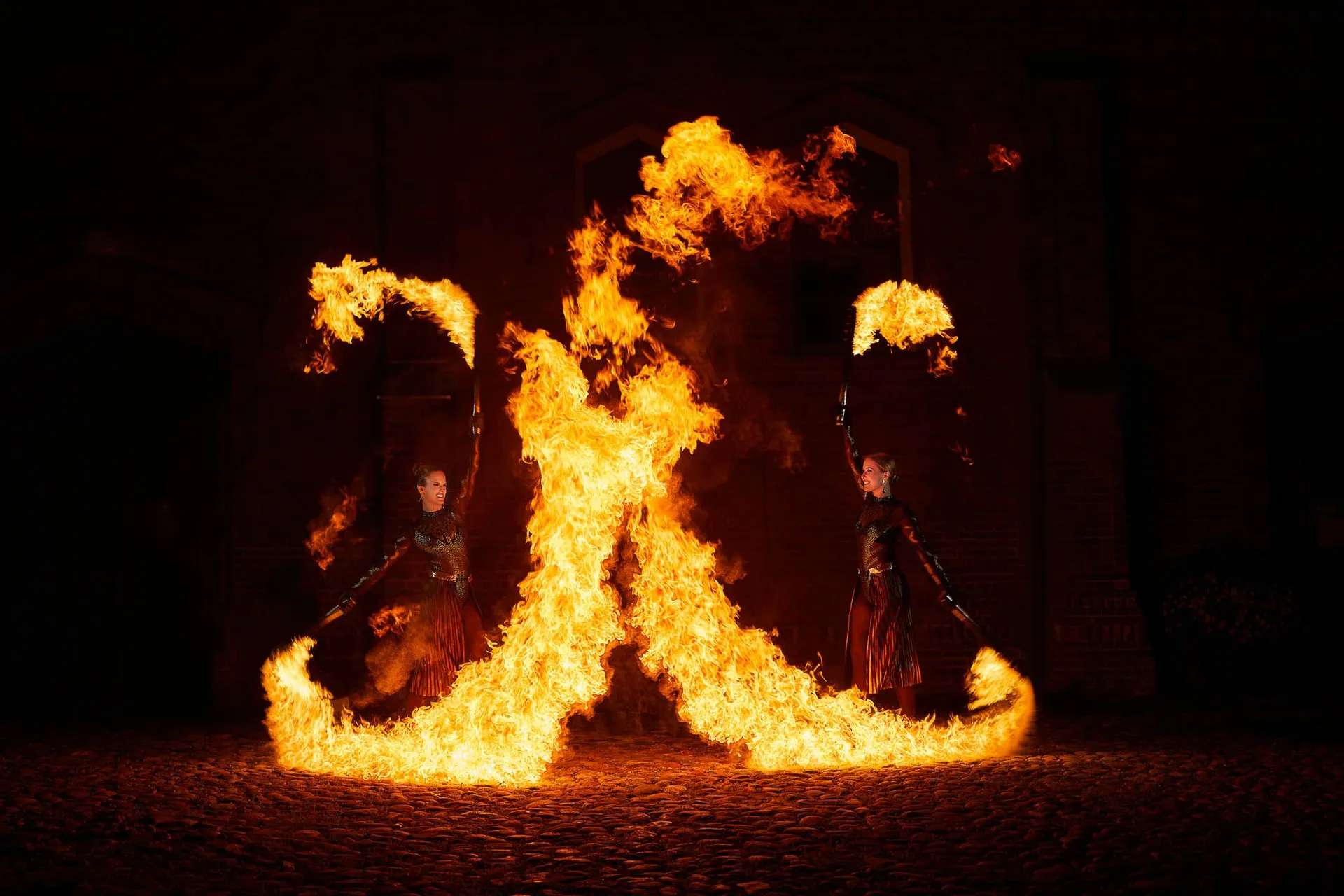 Two fire performers draw a large cross of flame in front of a brick building