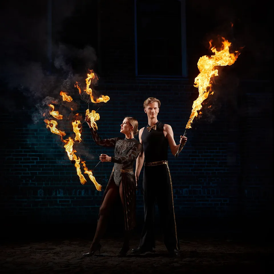 A duo by a brick wall at night: a dancer with fire fans and a performer holding a burning torch
